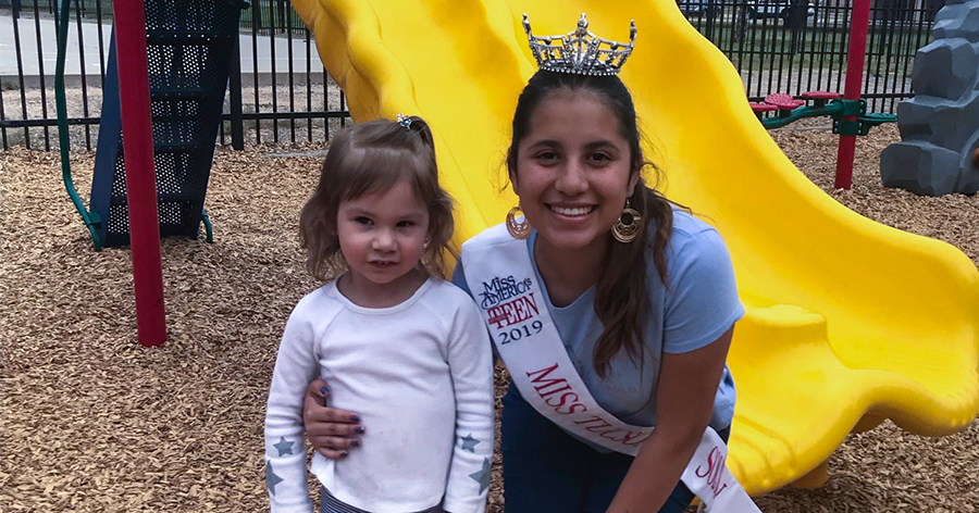 Miss City of Tucson Outstanding Teen is with a little girl in front of a slide at a playground.