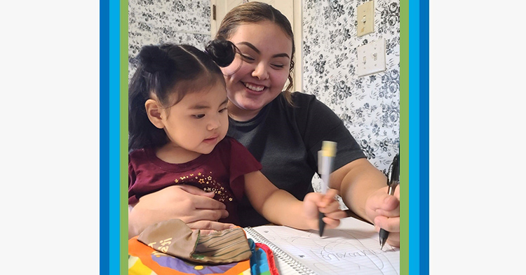 Mom with black tshirt holds her toddler daughter in her lap as they draw with markers.