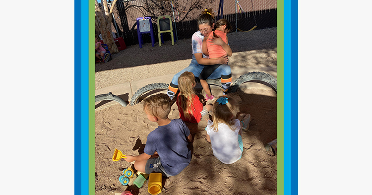 Woman in pink shirt cares for young children outside in a sandbox.