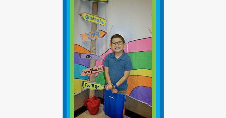 Young girl holding balloons, wearing a graduation cap in front of a preschool graduation poster