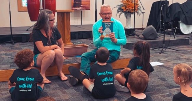 children sitting on church floor listening to a teacher read them a book.