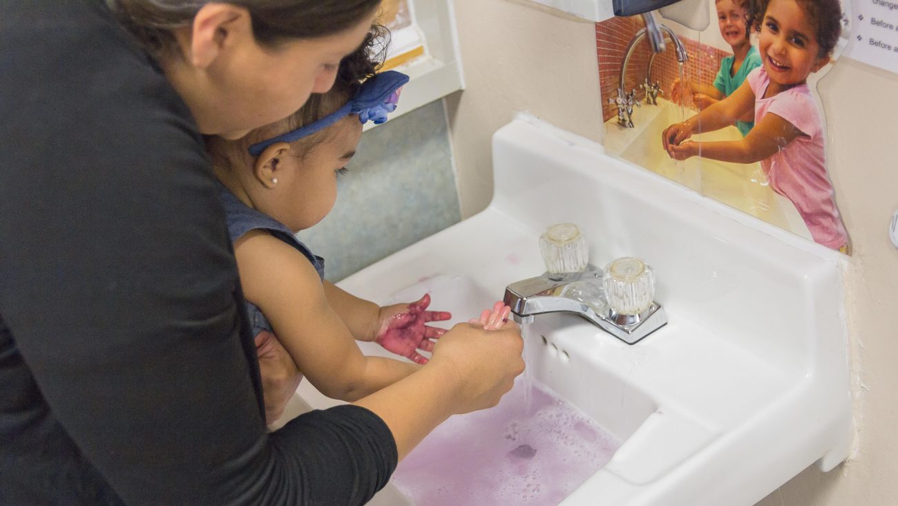 Teacher holding a young child over a sink helping to wash the child's hands.