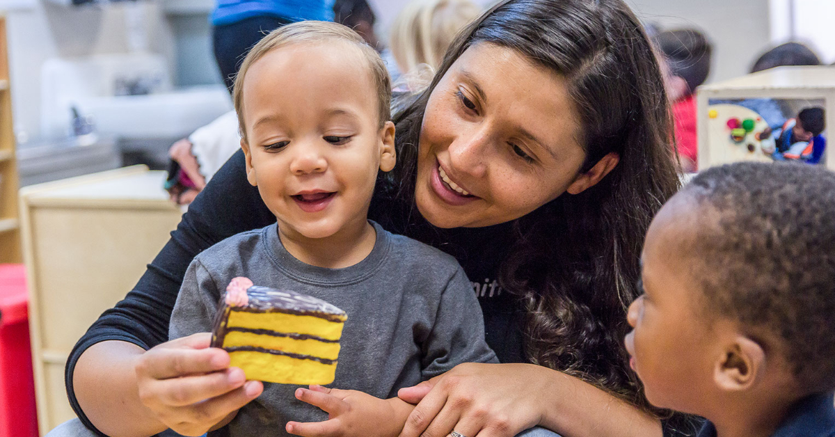 child care teacher holding little boy with a toy piece of cake.