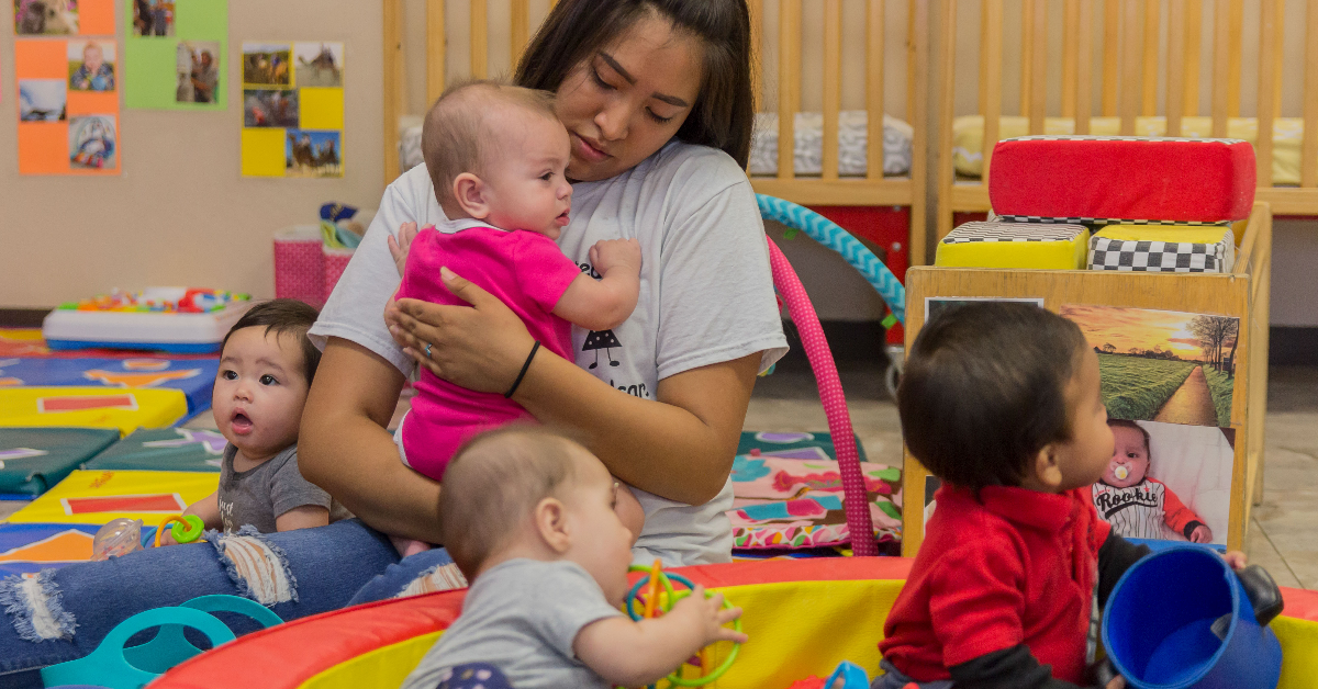 child care provider holding baby while two babies play on a mat.