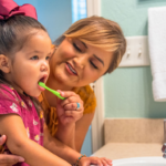 woman standing with young child at bathroom sink. Girl is brushing her baby teeth.