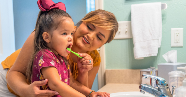 woman standing with young child at bathroom sink. Girl is brushing her baby teeth.