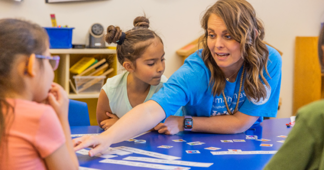 Preschool teacher reaching across a table with little children seated around her. Early childhood college scholarships