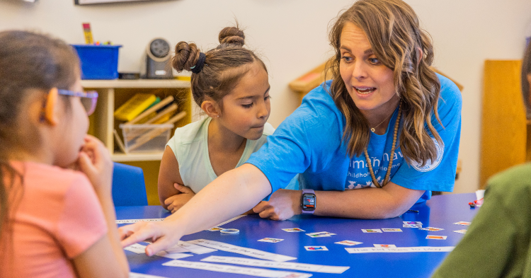 Preschool teacher reaching across a table with little children seated around her. Early childhood college scholarships