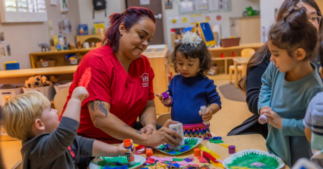 Woman wearing a red tshirt is in a classroom working with small chlidren gathered around a table with shapes. Child care shortages in Arizona.