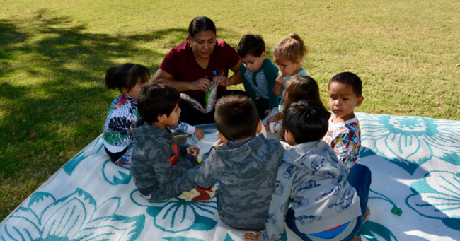 Preschoolers at Little Giants Spanish Immersion Preschool in Phoenix sit in a circle outdoors with teacher. 