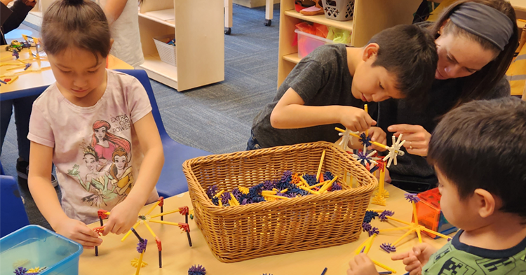 Three preschool-aged children around a small table playing with blocks. One child is sitting with an adult, who is helping him.