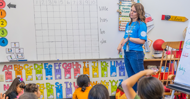 Preschool teacher standing in front of small children in a colorful classroom with the alphabet and white board on the wall.